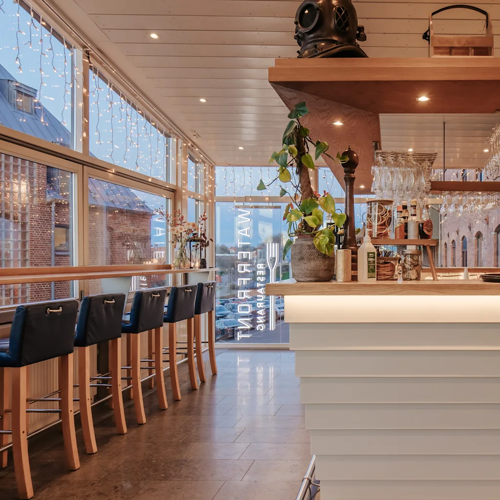 A modern bar area with a long counter, bar stools, and large windows. The bar features bottles, glasses, and a sign that reads WATERFRONT.