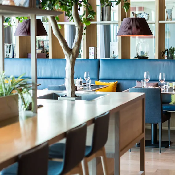 Bord dekket med glass og puter i en moderne restaurant, omgitt av innendørs planter og tretreinteriør.