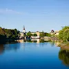 Rolig flod, der afspejler den klare blå himmel omgivet af frodige grønne træer og bygninger i baggrunden, herunder et fremtrædende kyrkespir under en solrig dag.