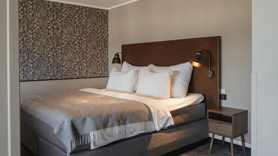 Et koselig hotellrom med en komfortabel seng, hvite sengetøy og et grått pledd, samt moderne vegglamper.