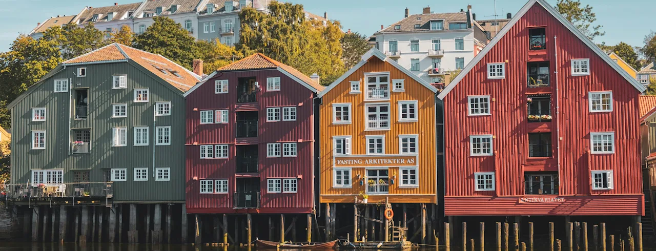 Värikkäitä puurakennuksia paalujen päällä tyynen veden yllä, teksti FASTING ARKITEKTER näkyvissä yhdessä. Kukkuloita taustalla.