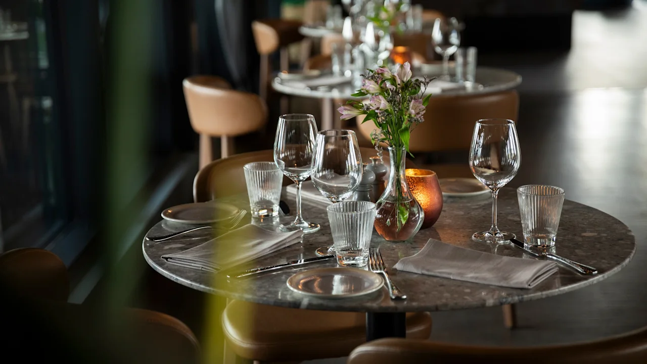 Et elegant dækket restaurantbord med vinglas, vandglas, bestik, servietter, blomster og et varmt lys.
