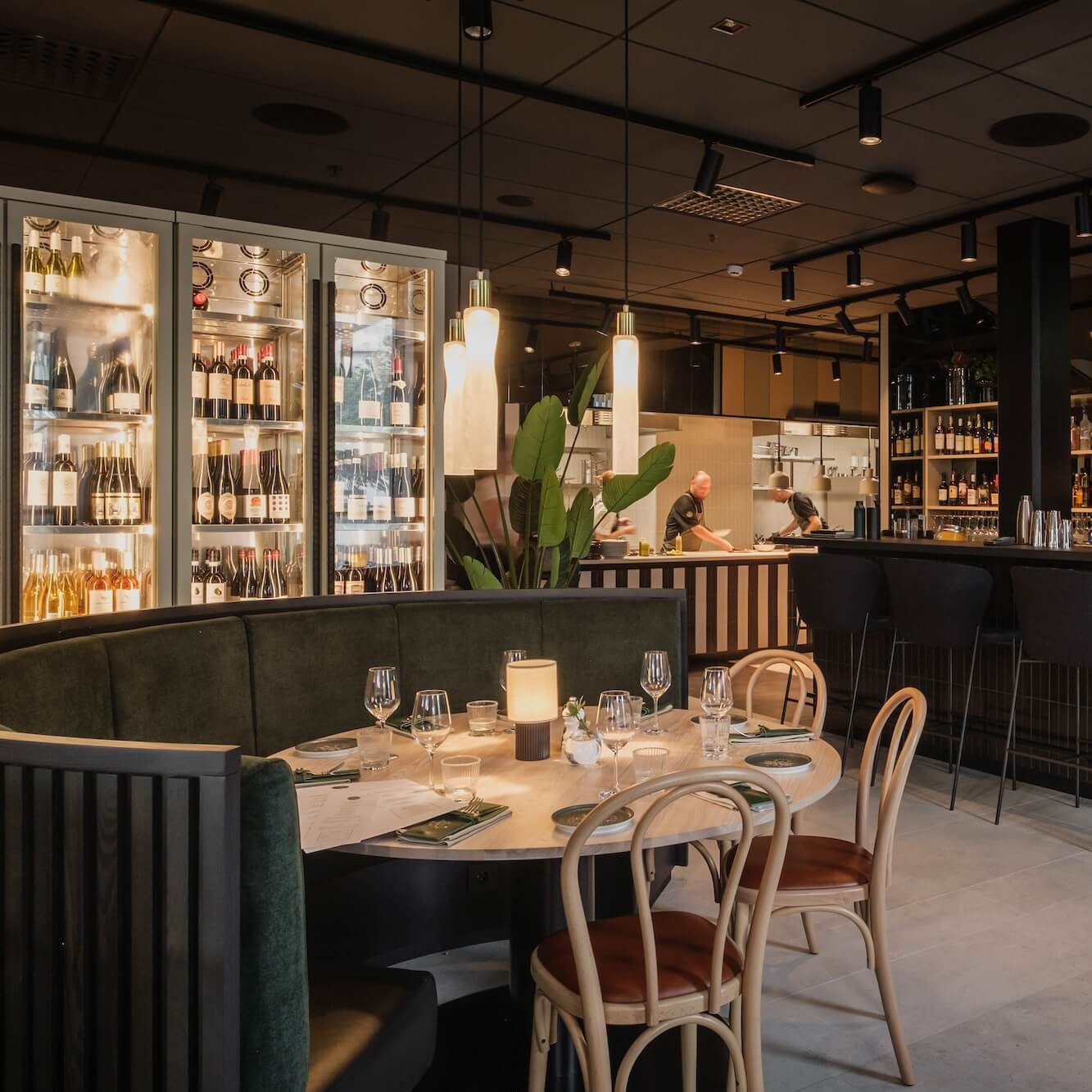 Elegant restaurantinteriør med gjester sittende ved bord og en bartender bak en bar, opplyst av pendellamper i moderne omgivelser.