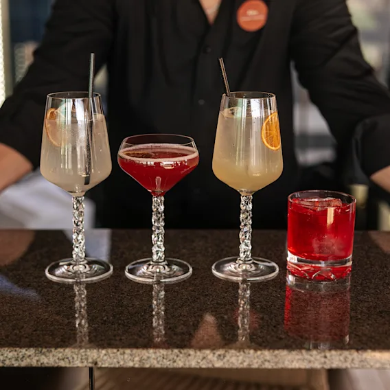 En bartender på Clarion Hotel Sign presenterar fyra färgglada cocktails på en bardisk, redo att serveras.