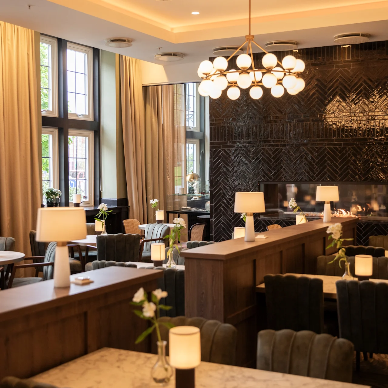Elegant dining room with marble tables and velvet chairs, softly illuminated by pendant lights and lamps. A modern fireplace enhances the sophisticated ambiance. Large windows offer natural light, framed by curtains.