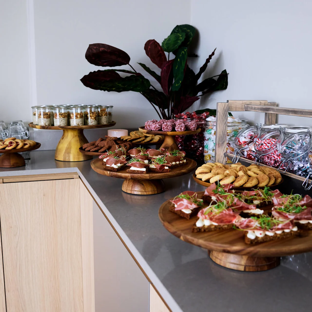 A wooden counter holds a buffet with assorted pastries, cookies, and jars of candies. Stacked glasses sit nearby, and decorative plants add greenery to the setup.