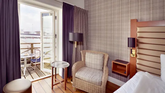 A cozy hotel room with a wicker chair and bedside lamp, opening to a balcony overlooking water.