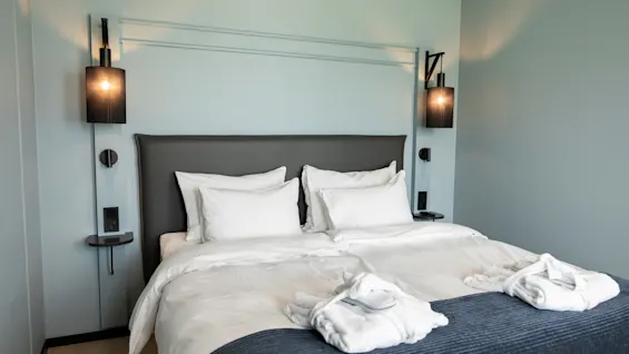 Et koselig hotellrom med en stor seng, hvitt sengetøy og to badekåper, med moderne vegglamper og en lyseblå vegg.