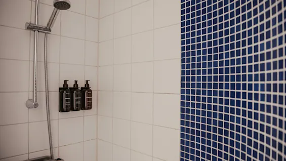 Moderni suihku Home Hotel Uppsalassa, jossa on valkoiset kaakeliseinät ja näyttävä sininen mosaiikkiaksentti, sekä kätevät seinäkiinnitteiset annostelijat.