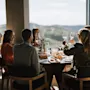 Folk sidder omkring et spisebord og løfter vinglas i en skål; store vinduer afslører en naturskøn udsigt over bakker under en klar himmel.