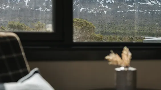 Utsikt över ett snötäckt berg genom ett fönster med en mysig fåtölj i förgrunden.