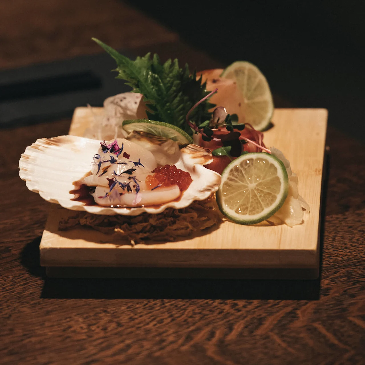 Sashimi presentert i et skjell på en treplate med skiver av lime og garnityr, rik på tekstur og farger.