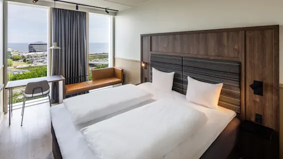 En pent dobbelseng står i et moderne hotellrom med store vinduer som gir utsikt over havet og nærliggende bygninger. En stol og et bord er ved siden av.