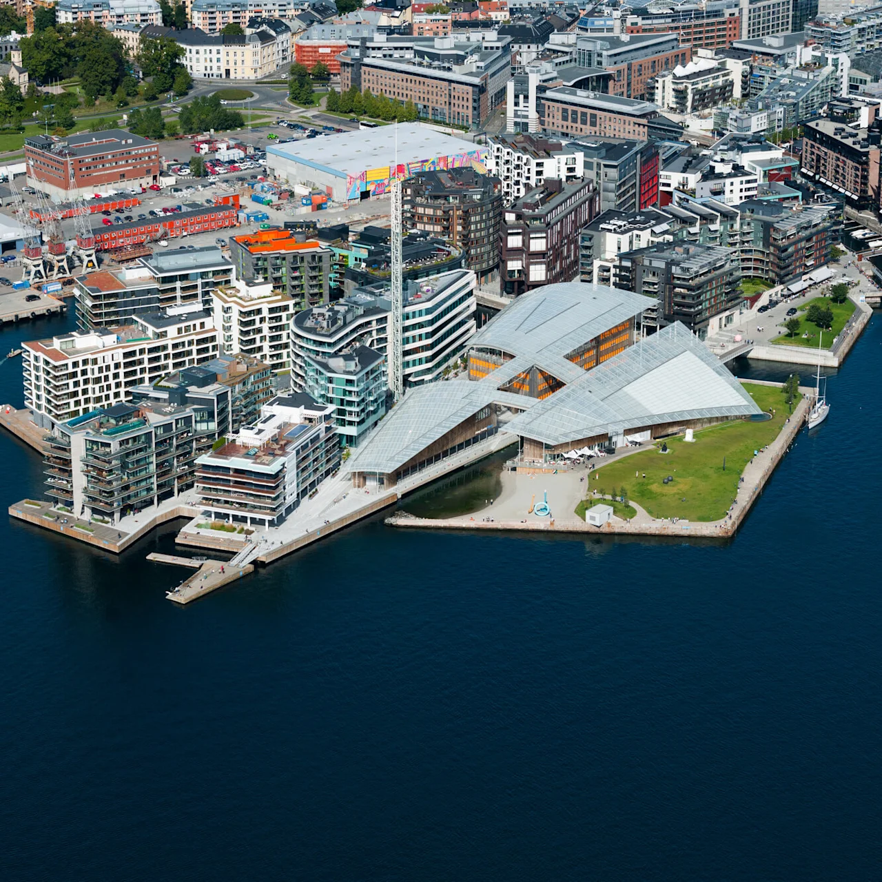 Moderne bygninger med unik arkitektur står langs en strandkant, omgitt av bystrukturer. Et historisk skip seiler i vannet i nærheten.