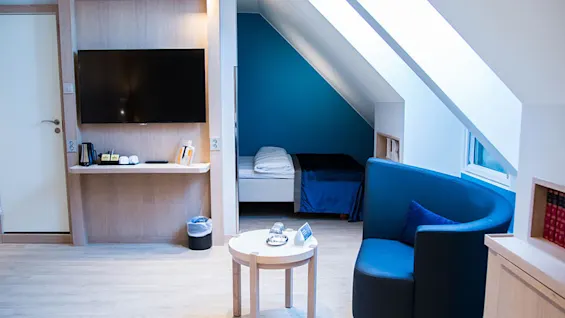 Et værelse med et vægmonteret TV, en enkeltseng under et skrånende loft, et lille rundt bord og en blå lænestol. Lysegulv og store skrå vinduer.