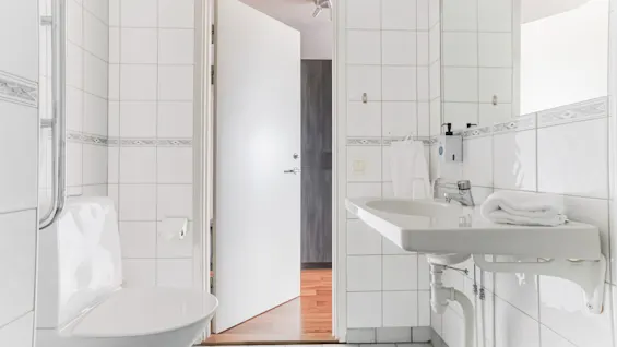 A clean, white-tiled bathroom with a toilet, sink, and mirror. An open door leads to another room.