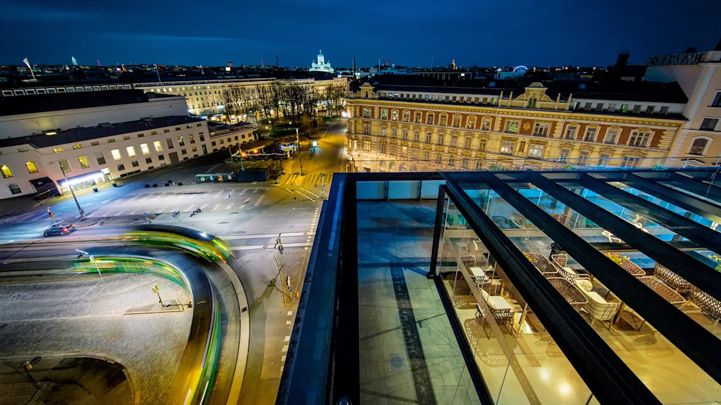 En trikk kjører langs en buet gate i et opplyst bylandskap om natten. Takterrasse med glassrekkverk har utsikt over gaten og omkringliggende bygninger.