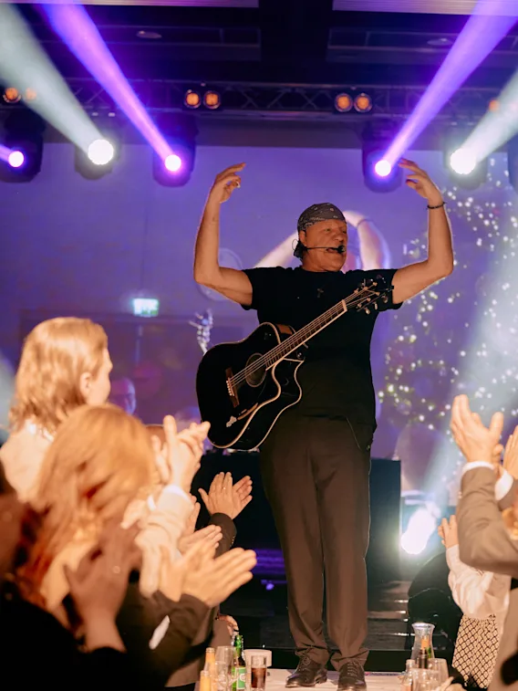 En musiker med gitar opptrer på en lyst opplyst scene, med armene hevet, mens publikum klapper og konfetti faller.