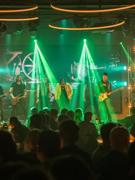 A band plays onstage, illuminated by vibrant green lights, in a crowded indoor concert venue with an audience seated at tables. Musical instruments and equipment are visible.