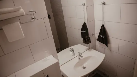 Kylpyhuone, jonka valkoinen allas ja sen yläpuolella oleva peili, kehystävät mustat "AQUAY"-tuotemerkin toilettilaukut koukuissa. Laatoitetut seinät ja pyyheteline antavat kontekstin.