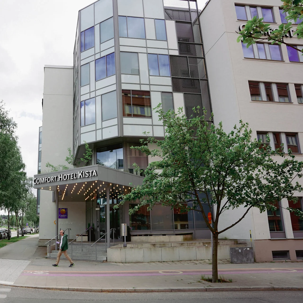 Person exiting the Comfort Hotel Kista, trees and cars present, urban street setting.