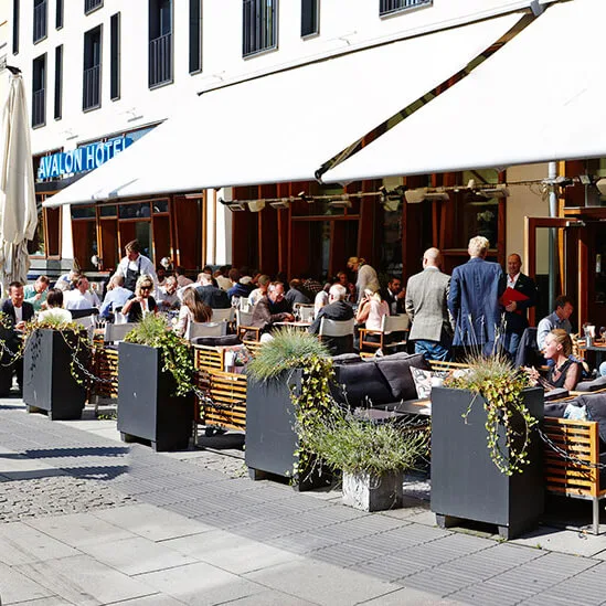 Outdoor café with people sitting at tables, eating and socializing under a white awning. Surrounded by large planters with greenery. "AVALON HOTEL" sign visible on the building.