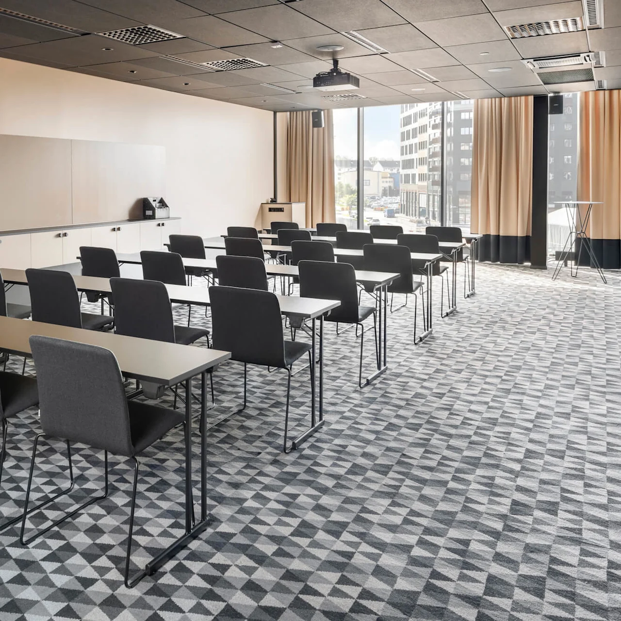 Svarta stolar i rader vända mot långa bord i ett konferensrum med geometrisk matta, projektor, stora fönster och beige gardiner.