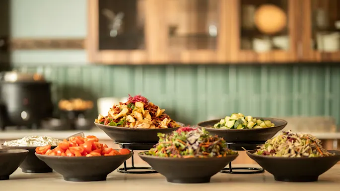 Boller fylt med ulike fargerike salater og grønnsaker vises på en trebord i et kjøkken med grønne flisvegger.