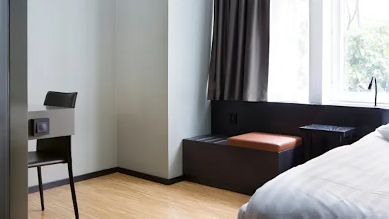 Stol ved siden af et skrivebord står i et minimalistisk rum med lyst trægulv, ved siden af en seng og et vindue med mørke gardiner. En sort bænk og et lille bord er i nærheden.