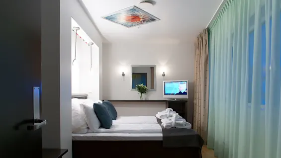 A neatly made bed with pillows and towels, a mounted TV displaying a news channel, in a clean, well-lit hotel room with curtains.