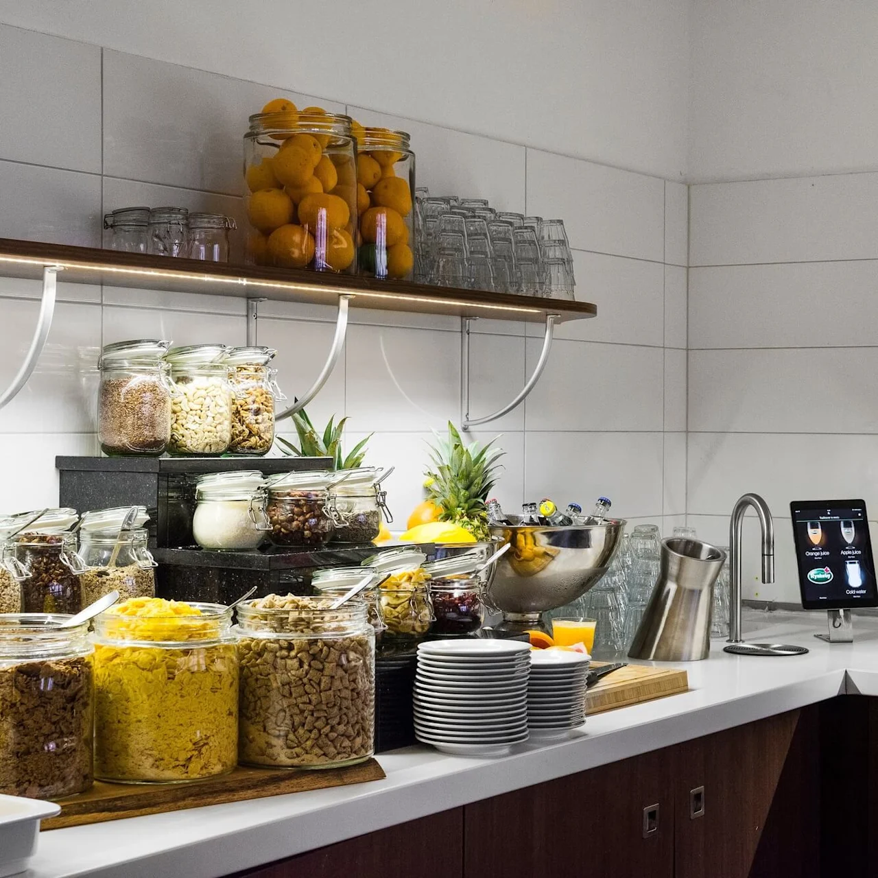En moderne buffet med ulike frokostblandinger i glasskrukker og fruktboller på en benk, satt mot en flislagt vegg med hyller.