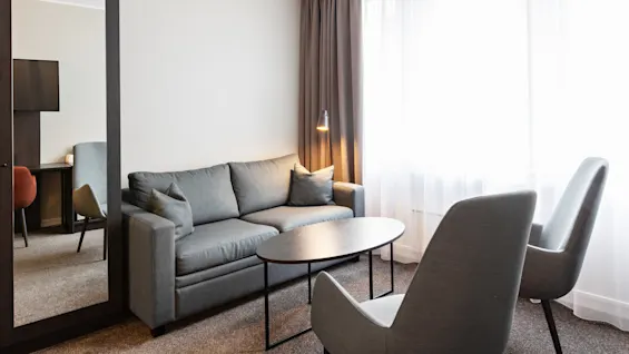 En grå sofa står i stuen med to stole og et sort sofabord. En gulvlampe oplyser rummet ved siden af ​​lukkede gardiner, med et vægspejl, der reflekterer en del af et skrivebord.