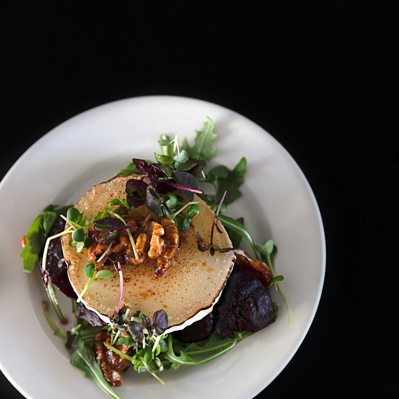 En salat står på en hvit tallerken, toppet med blandede grønnsaker, pæreskiver, valnøtter og muligens en balsamico-skvett, sett mot en svart bakgrunn.