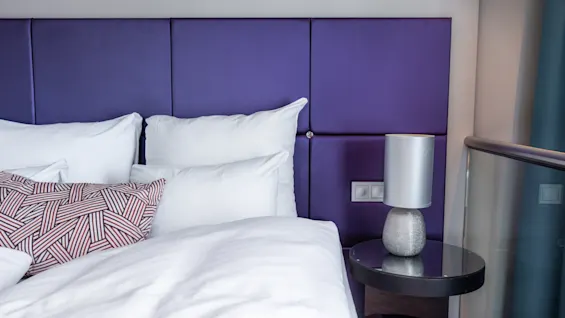 Bed with white pillows and sheets, resting against a purple padded headboard, next to a round bedside table holding a modern lamp. Brown comforter and patterned pillow add contrast.