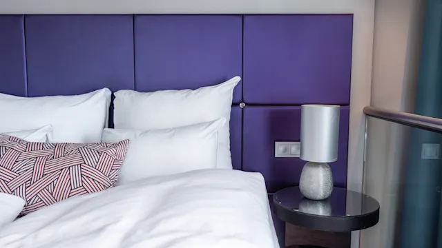 Bed with white pillows and sheets, resting against a purple padded headboard, next to a round bedside table holding a modern lamp. Brown comforter and patterned pillow add contrast.