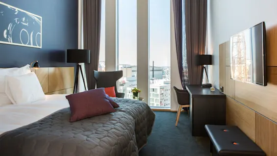 Et moderne hotellrom med en pent oppredd seng med puter, et arbeidsbord og et stort vindu med byutsikt. Nøytrale toner og minimalistisk innredning.