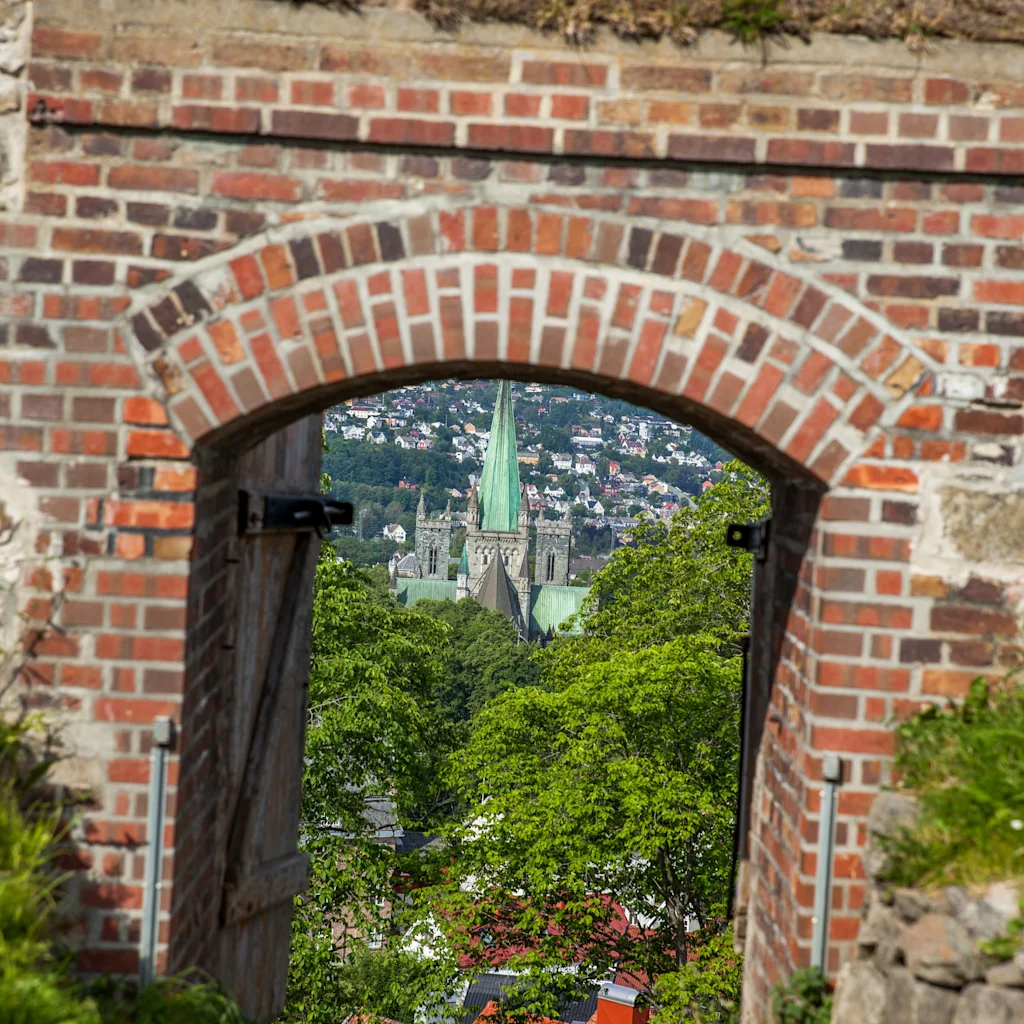Utsikt gjennom en gammel mursteinsbue som avslører en by med en stor kirkebygning og grønne trær under en lys himmel.