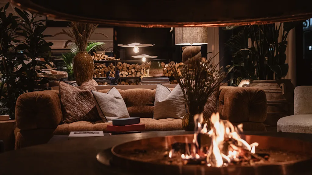 Ildsted flammes, omgitt av en koselig brun sofa med puter. Varmt lys fra lamper forsterker det rustikke miljøet, med planter og vedkubber i bakgrunnen.