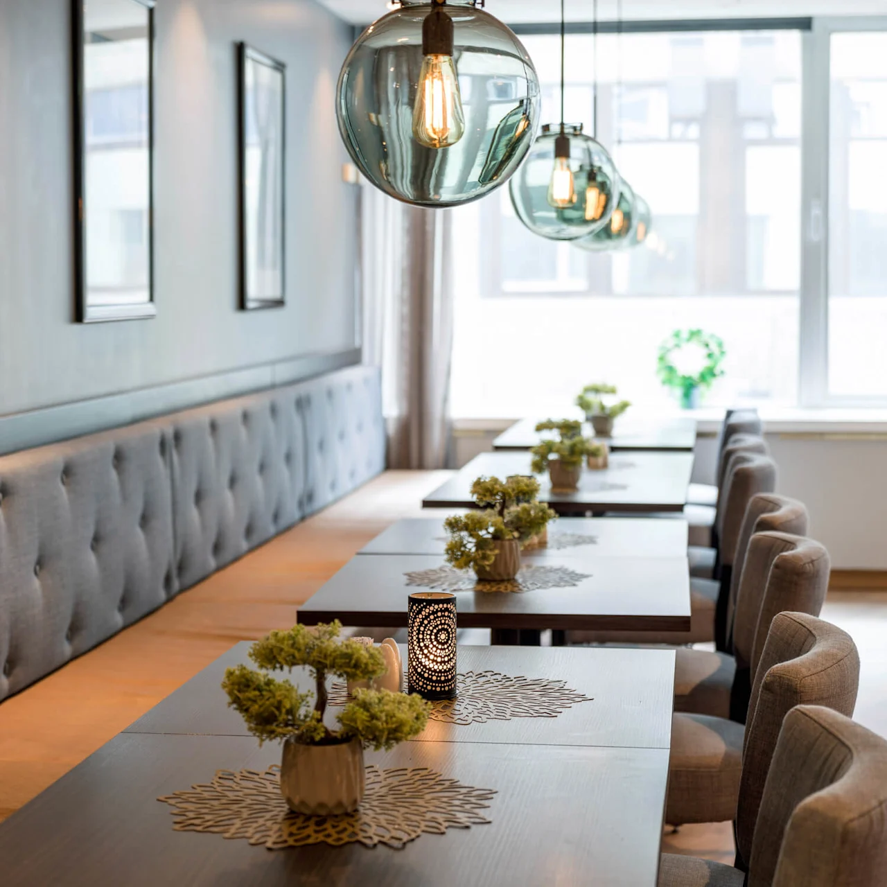 Modern dining table adorned with small potted plants on stylish mats, surrounded by plush chairs. Hanging globe lights illuminate the bright restaurant interior alongside large windows and framed wall art.