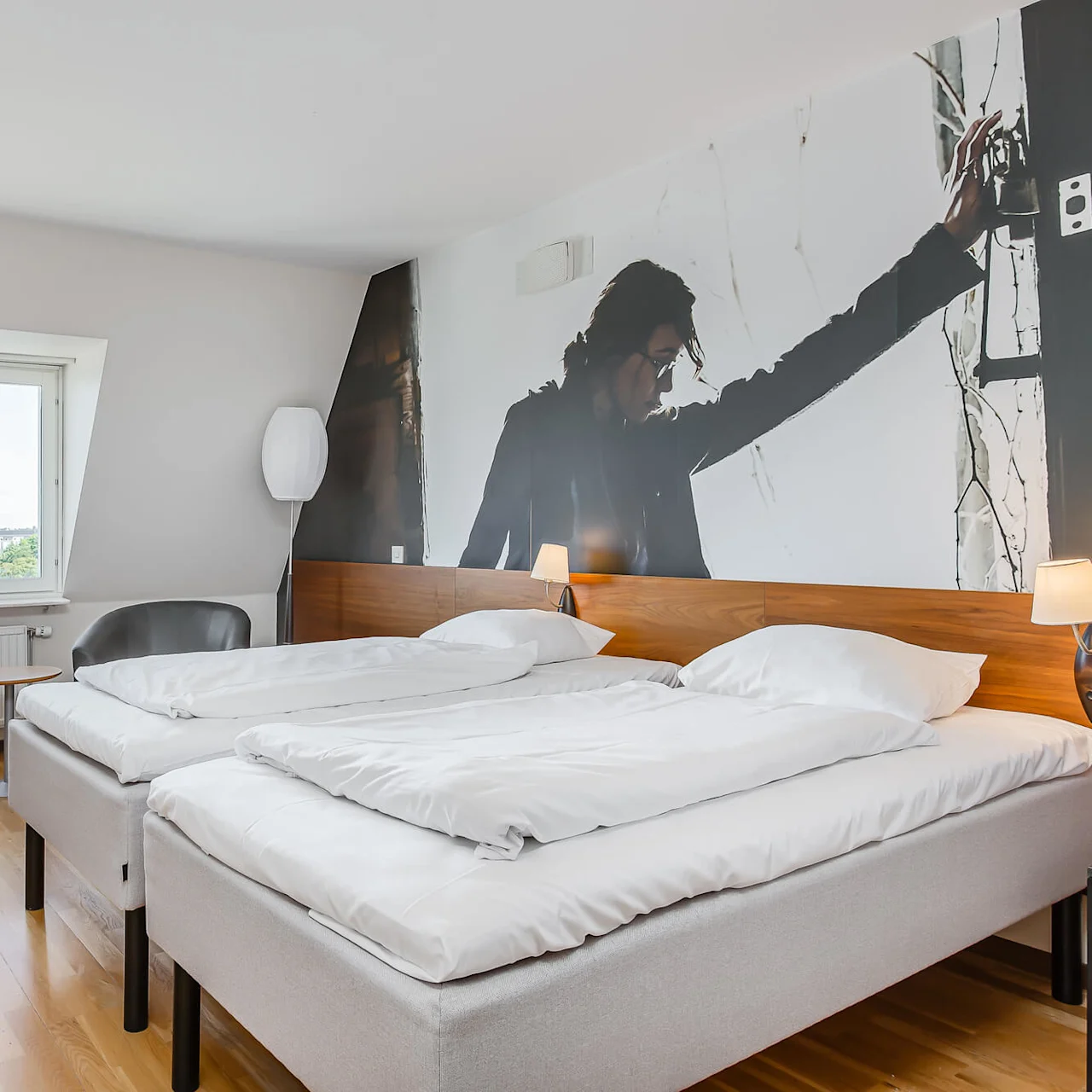 Hotellihuoneessa on kaksi siististi pedattua sänkyä, suuri seinämaalaus takkiin pukeutuneesta henkilöstä, kirjoituspöytä tuolin kanssa ja ikkuna, josta näkyy vihreää maisemaa ulkopuolella.