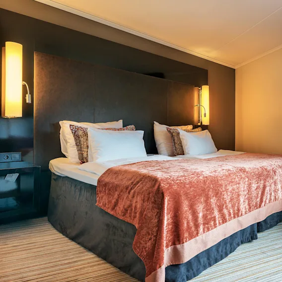 A neatly made bed with white pillows and a peach blanket rests against a dark headboard, flanked by wall lamps, beside a window with brown curtains in a carpeted room.