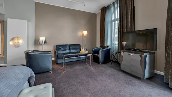 En stue har en sort lædersofa placeret omkring et glasbord, en gulvlampe og en stor fladskærms TV på et skab. Brune gardiner dækker vinduet.