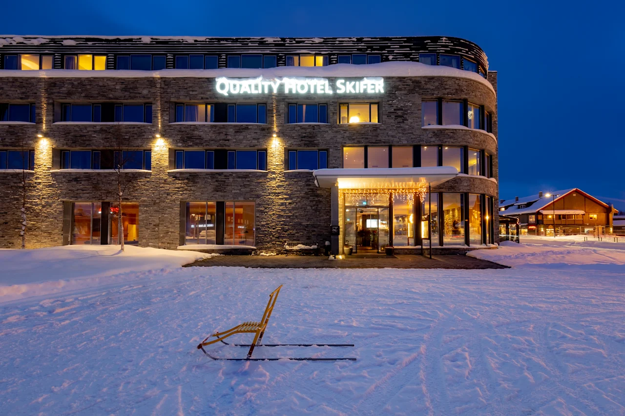 En kjelke står på snøen foran en fleretasje bygning med opplyste vinduer, navngitt "QUALITY HOTEL SKIFER." Det omkringliggende lyset skaper en koselig vinterkveldstemning.
