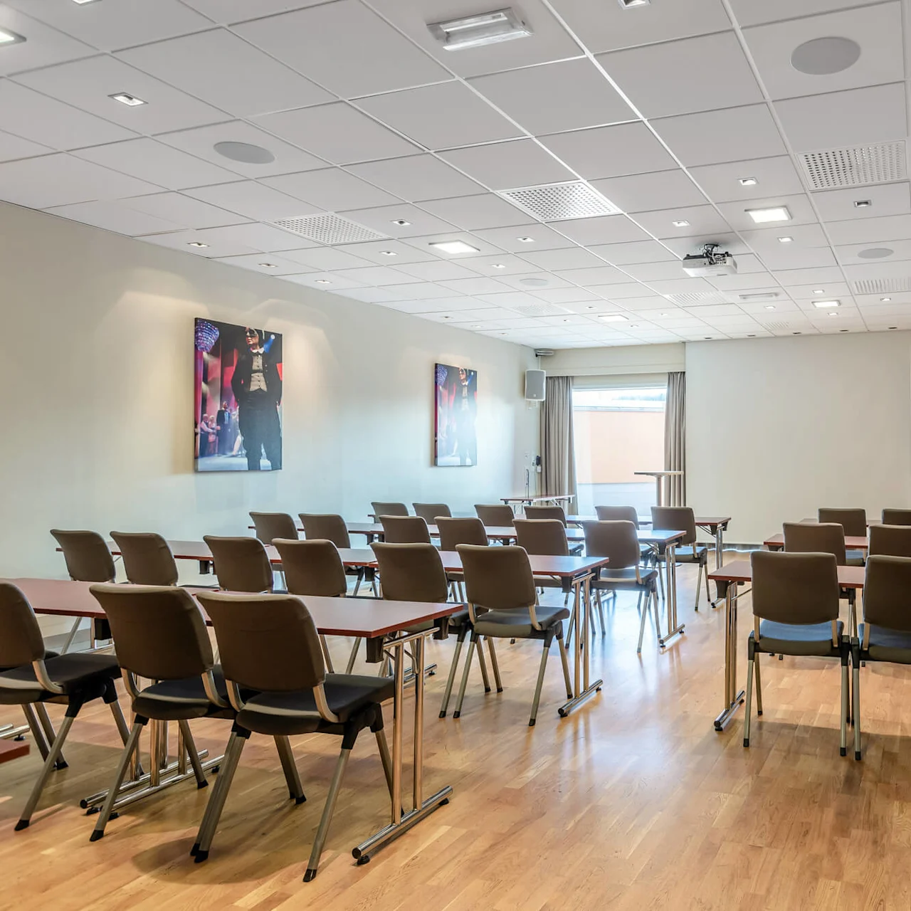 Rader med tomme stoler vender mot et presentasjonsområde med en whiteboard i et konferanserom med tregulv og malerier på veggene.