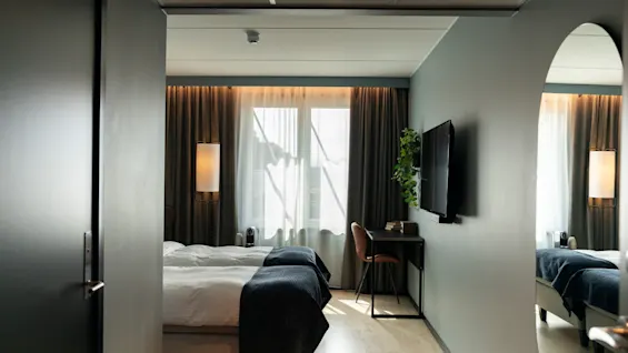 Et moderne hotellrom med to senger, et skrivebord, en TV og et stort vindu med gardiner.