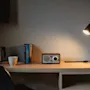 Ett träskrivbord har en retro radio bredvid en tänd svart lampa. I närheten står en vattenkokare och en mugg, med böcker prydligt uppradade i en modern kontorsmiljö.