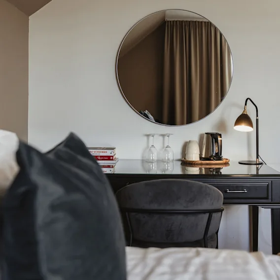 Et hotellrom med et skrivebord, stol og et speil. Bøker N SCHULMAN og Tage, en vannkoker og glass står på skrivebordet.