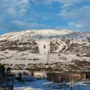 Snødekte fjellskråninger med skiløyper strekker seg ned mot en landsby med spredte hus og bygninger, omgitt av skoger under en delvis skyet blå himmel.