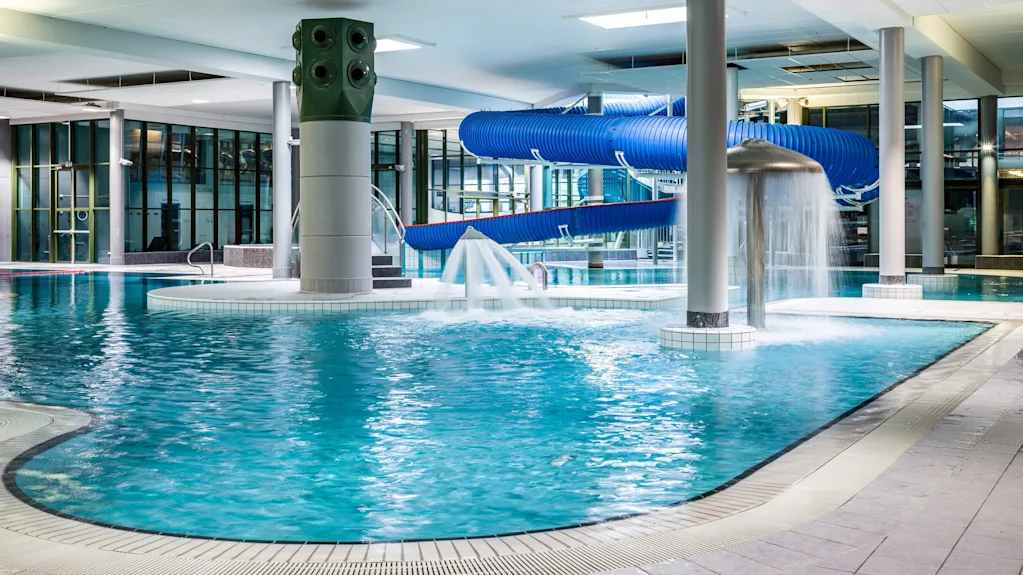 Inomhuspool med blå vattenrutschbana och fontän; omgiven av kakelgolv och stora fönster. Ingen text finns.