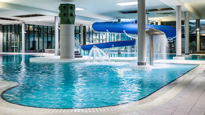 Indoor pool with blue water slide and water fountain; surrounded by tiled flooring and large windows. No text present.